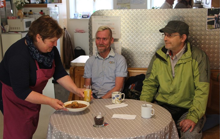 Wärmestube Schongau - An einem gedeckten Tisch mit umlaufender Bank sitzen 2 Personen und eine Wärmestuben-Mitarbeiterin bringt diesen Personen ein Mittagessen an den Tisch.
